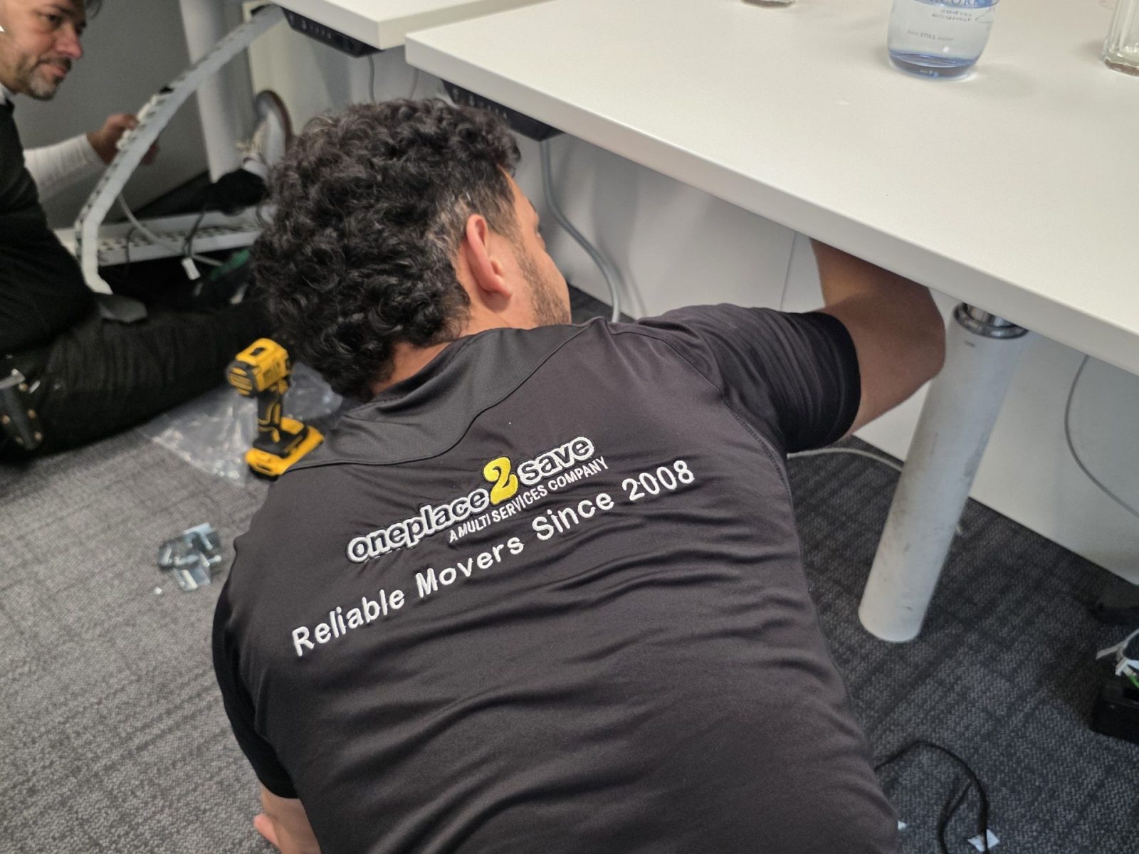 Oneplace2save team dismantling a white office desk on-site, disconnecting cables and preparing furniture for removal.